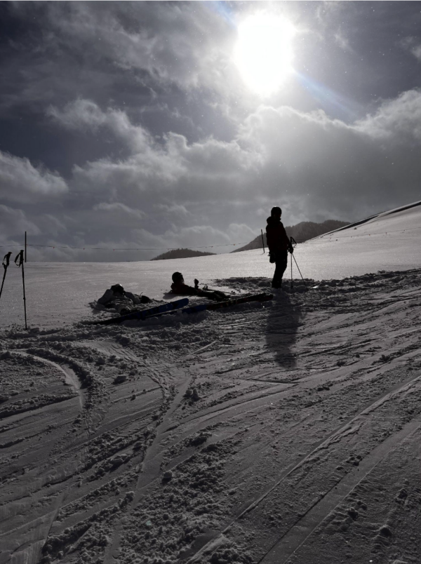 A person with ski poles stands on a snowy slope beside equipment and another person lying down, enjoying the bright sun—perhaps members of a local ski and snowboard club.